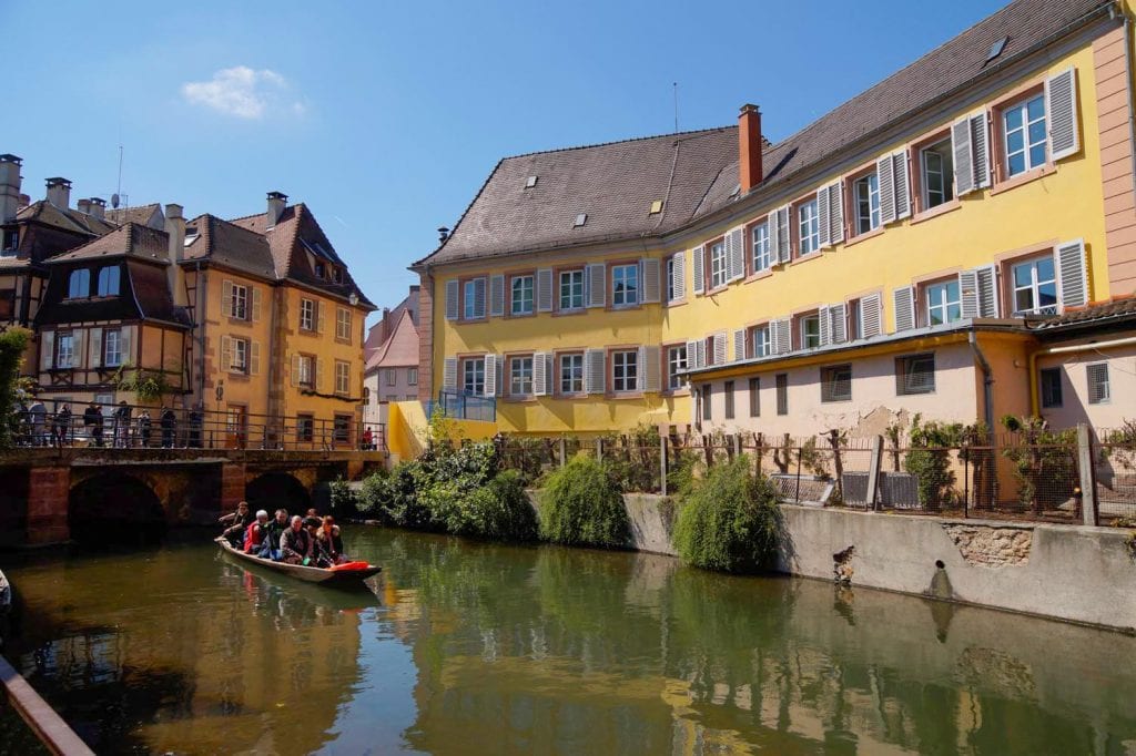 motorbootje vol met mensen in een kanaaltje in Colmar. Erachter lopen mensen over de brug en zie je vakwerkhuizen.