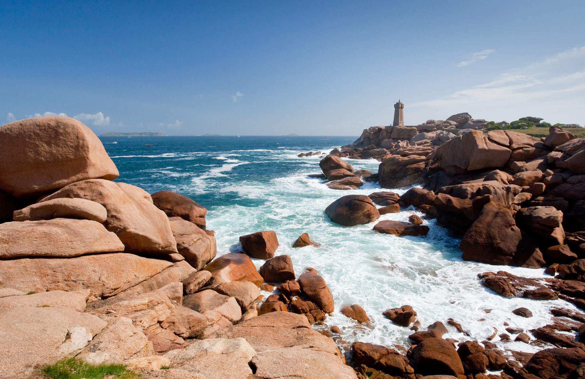 Roze granietrotsen en helderblauw water met in de verte een vuurtoren, de Côte de Granit in Bretagne. 