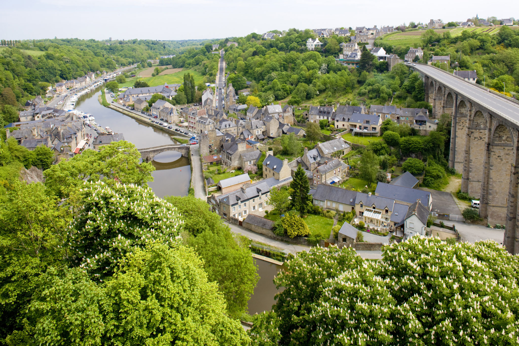 Luchtfoto van de middeleeuwse stad Dinan met huizen omringd door een rivier, veel bossen en een stenen brug. 