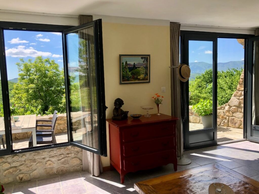 woonkamer van een vakantiewoning in Val-les-Bains met grote raampartijen en uitzicht op een terras en de beboste heuvels in de Ardèche