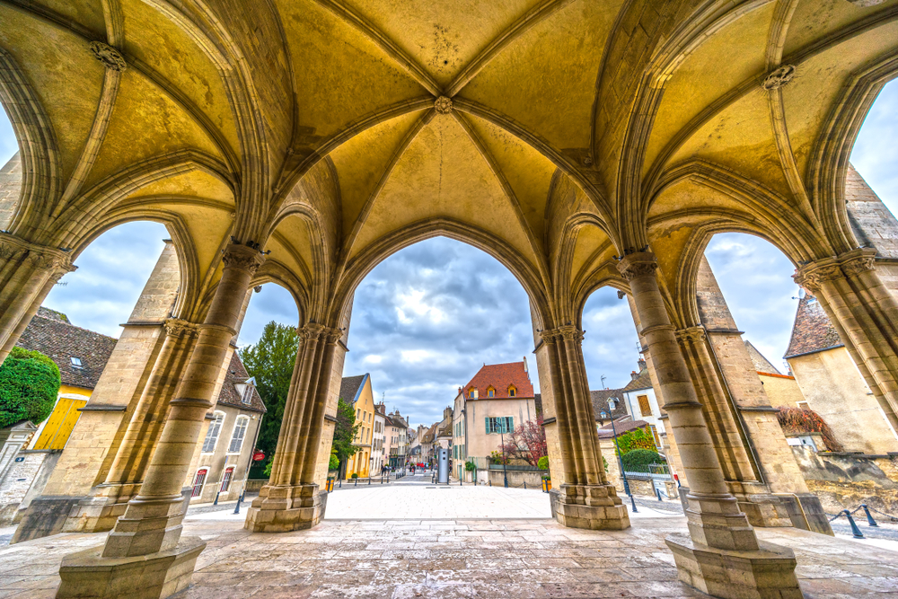 foto genomen van onder de stenen bogen van de Église de Notre-Dame in Beaune met zicht op een plein, gekleurde huizen en een straat 