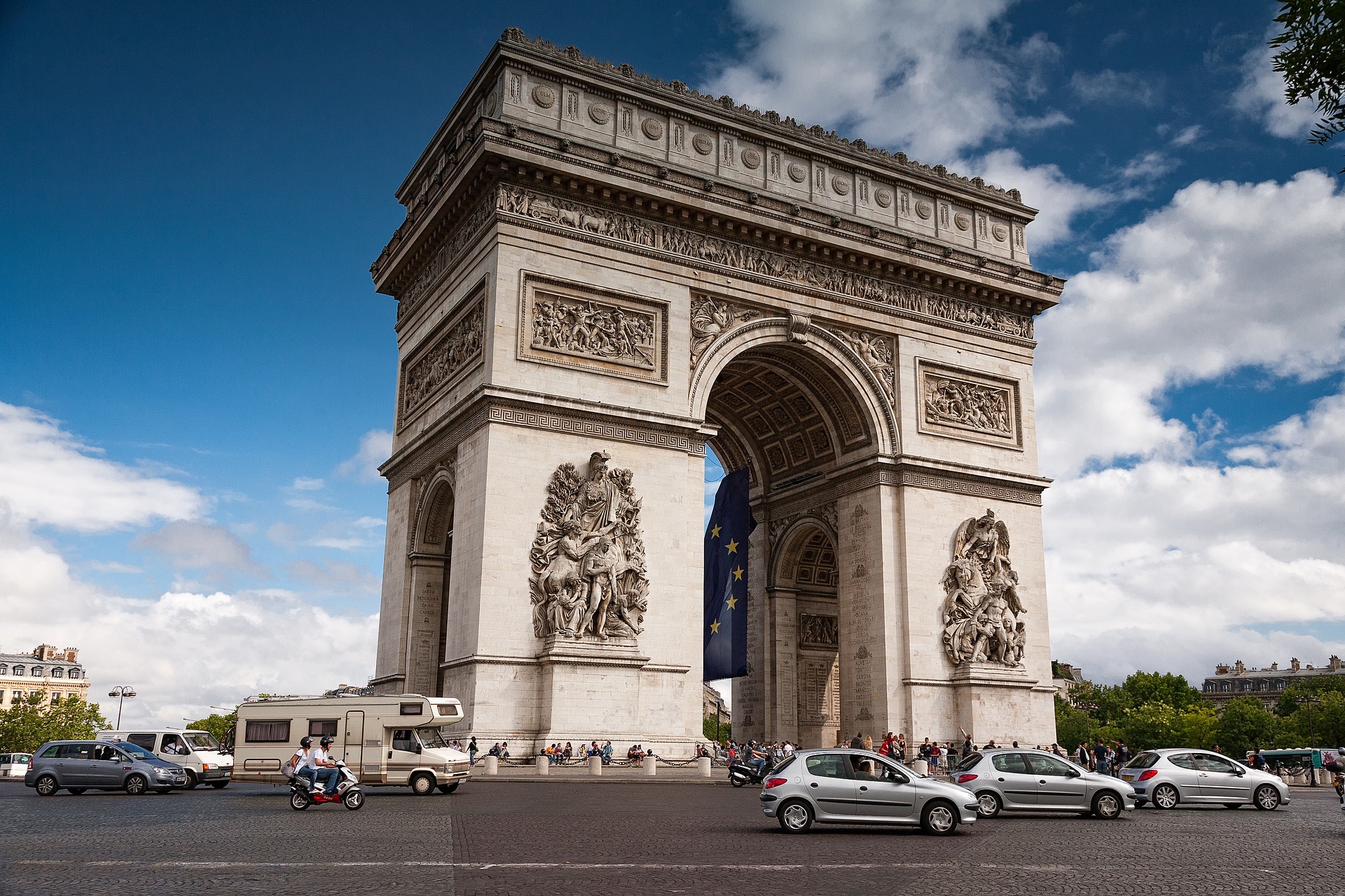 de Arc de Triompe met daarvoor grijze auto's, een camper, scooter en een wit busje die op de rotonde rijden