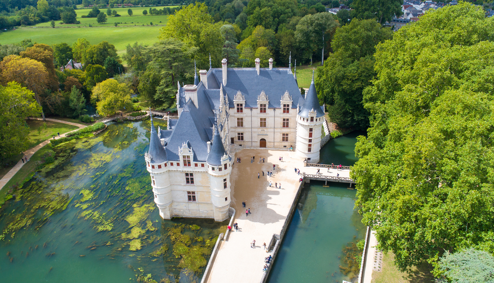 Wit kasteel met grijze puntige daken gelegen midden in het water en omringd door veel groene bomen en bossen.