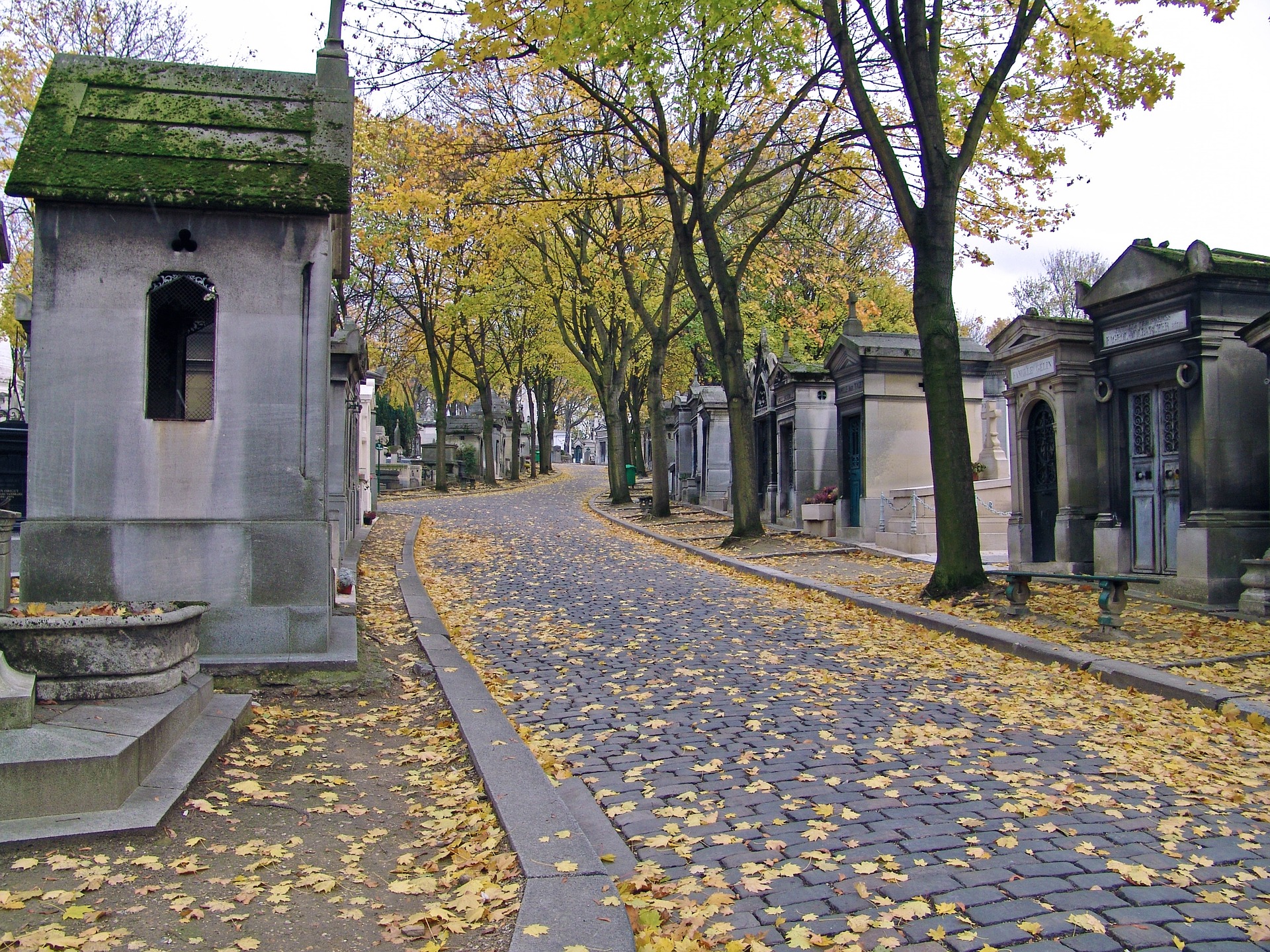 grafhuisjes op de begraafplaats Père Lachaise. Je kunt zien dat het herfst is, want er liggen en hangen gele blaadjes aan de bomen.