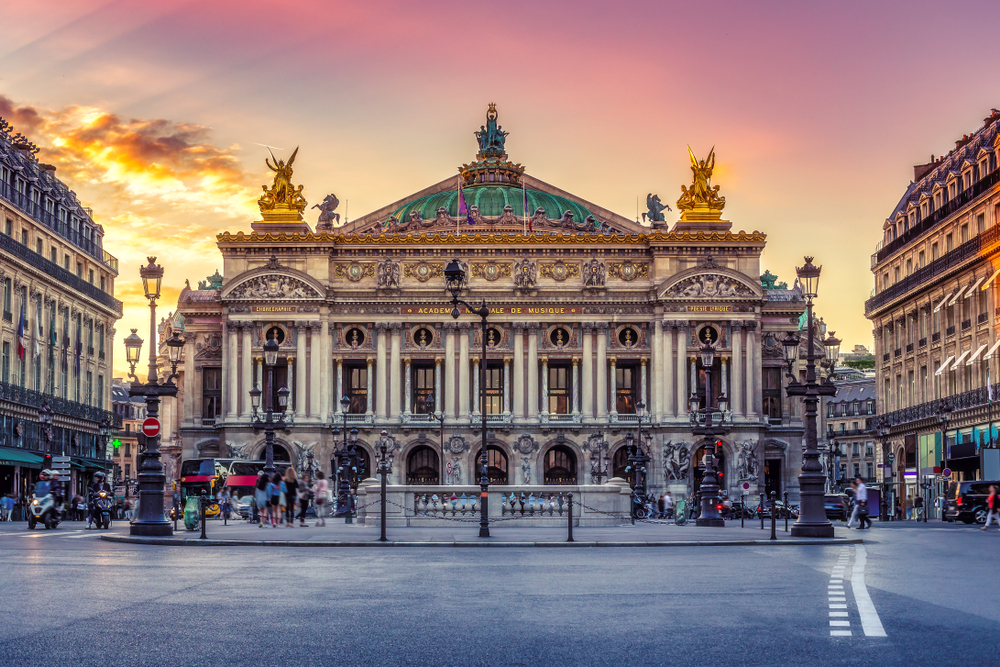 het operahuis Opéra Garnier in Parijs tijdens ondergaande zon De lucht kleurt roze/paars en er is wat verkeer op de straten.