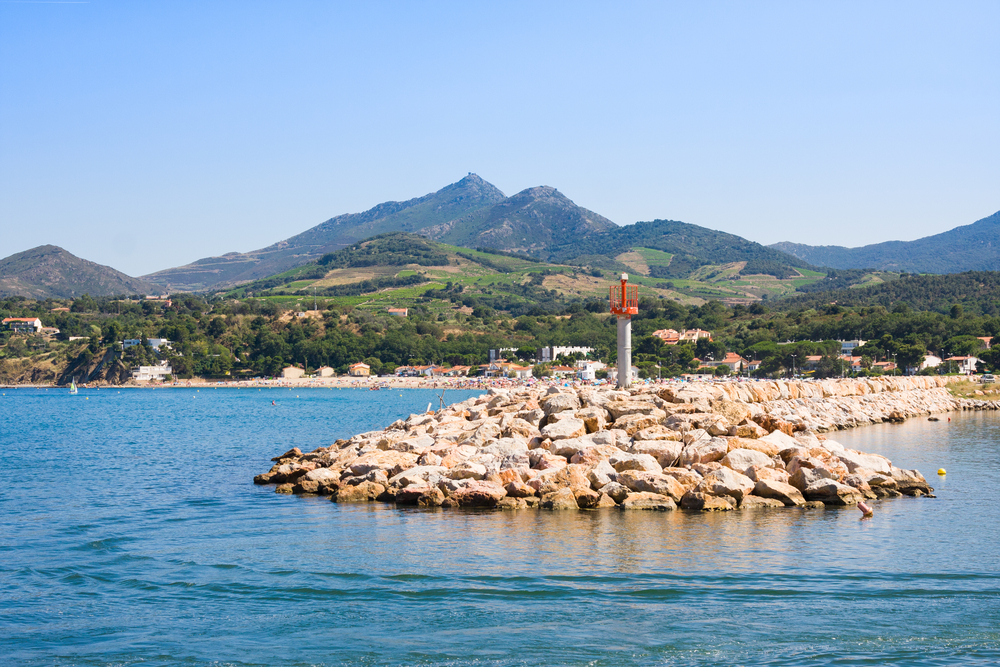 een stenen pier in Argeles sur Mer met op de achtergrond een aantal huisjes en daarachter groene heuvels