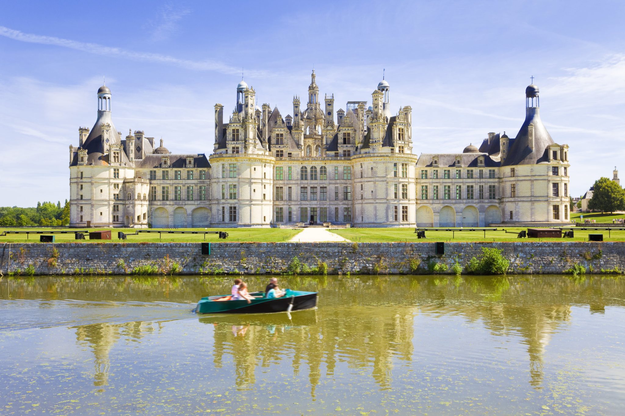 Groot wit kasteel met veel verschillende torentjes met zwarte daken gelegen op grasland aan rivier de Loire waar een bootje met mensen overheen varen.