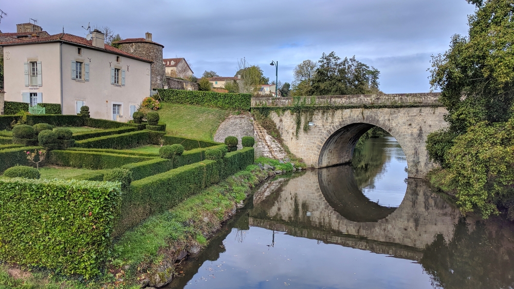 Stenen brug over rivier de Mère met huisjes en struiken aan de overkant.