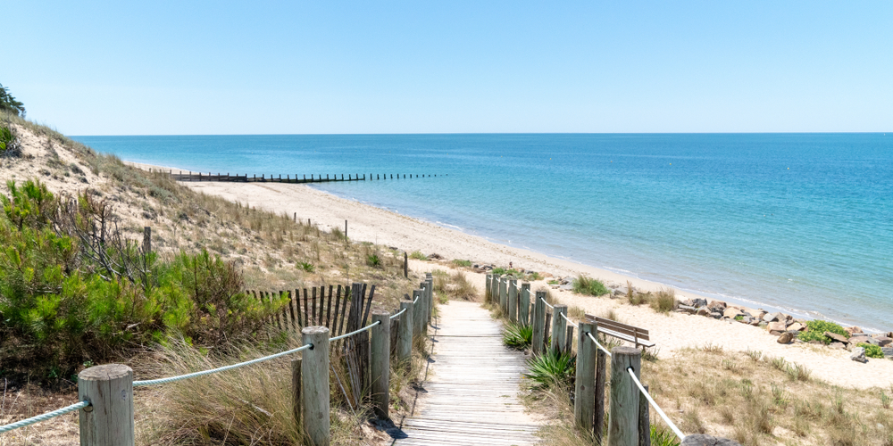 houten paadje met aan beide kanten palen die met touw verbonden worden en dat afloopt naar een zandstrand met azuurblauwe zee op het Île de Noirmoutier