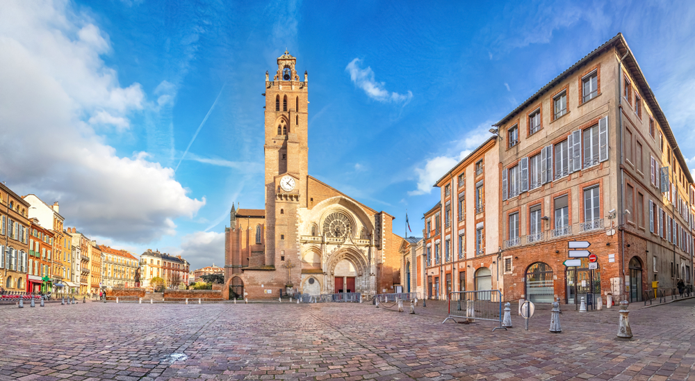 leeg plein met aan de zijkanten roze en gele gebouwen en een kerk met een hoge toren in Toulouse