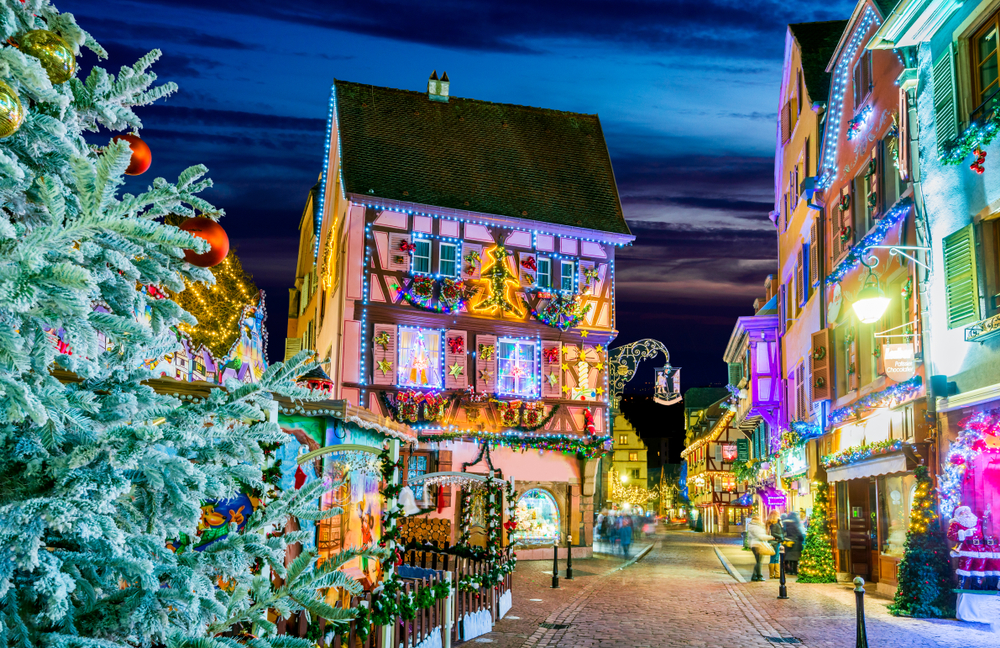 Huizen met kersversiering in een straat in Colmar met op de voorgrond aan de linkerkant een deel van een kerstboom. Er lopen enkele mensen op straat die door de ramen van winkels naar binnen kijken.