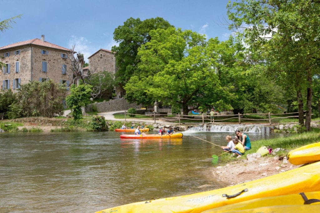 twee kano's met mensen op een rivier en kinderen die aan de kant aan het vissen zijn. Op de voorgrond liggen gele kano's