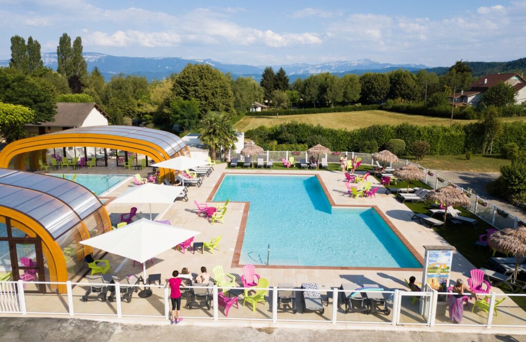 foto van de zwembaden op Camping Le Coin Tanquille met rondom ligbedden en roze en groene stoelen. Op de achtegrond zie je veel groen en daarachter weer de bergen.