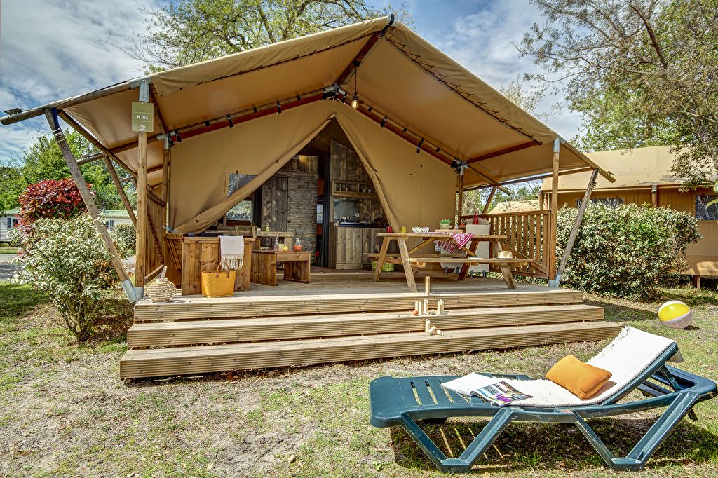 sfeervolle safaritent die open staat met een inrichting van veel hout en een gedekt terras. op de voorgrond zie je een zonnebedje met handdoek, kussen en tijdschrift