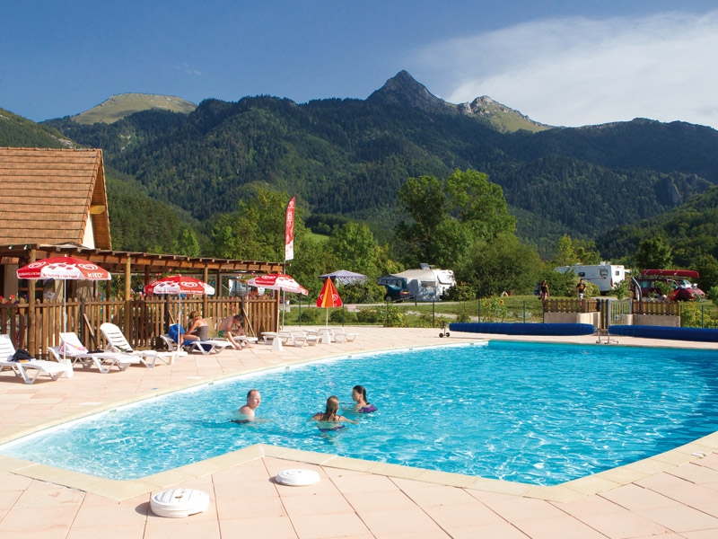 zwembad met daarin drie mensen en aan de kant een moeder en een kind op ligstoelen op Camping Sites et Paysages Belle Roche. Op de achtegrond zie je groene bergen en tussen de ligstoelen staan Ola-parasols