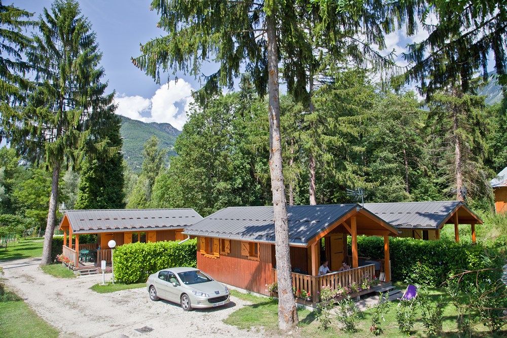 houten chalet op en camping tussen de bergen en met veel bomen. Op het terras van één van de twee chalets zit een stel aan de eettafel