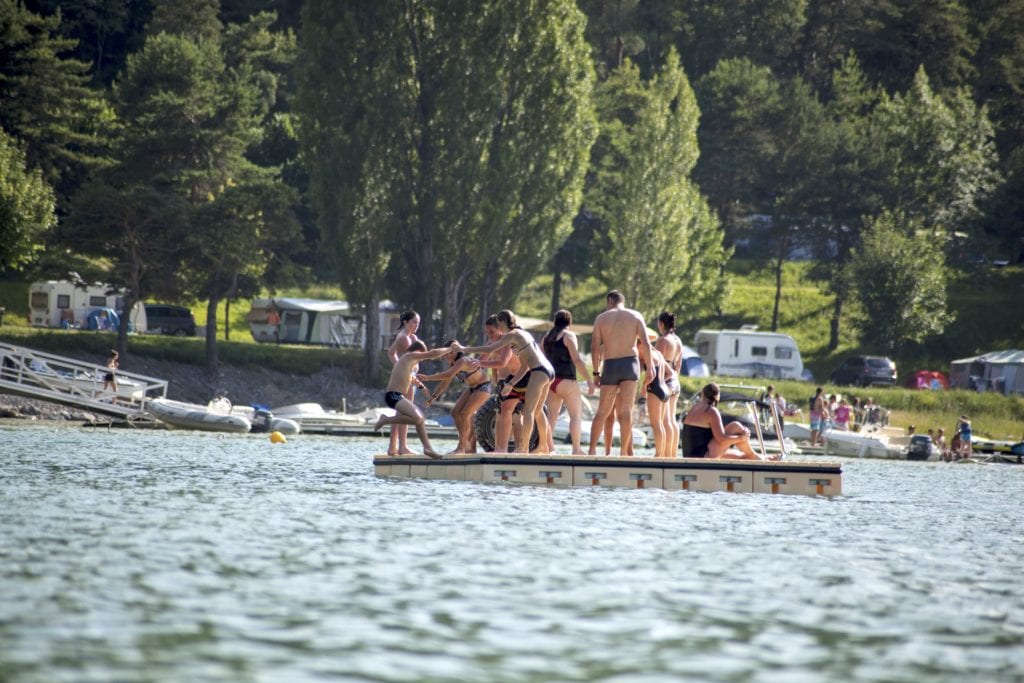 een man en kinderen op een vlot in een meer die elkaar van het vlot afduwen. Op de achtergrond zie je een camping met caravans liggen.