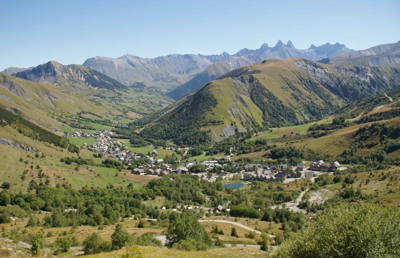 een groen dal met op de achtergrond bergen. In het dal ligt een meertje en een langgerekt dorp