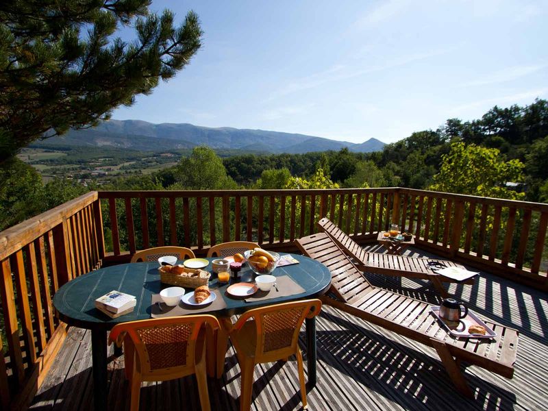 het terras van een chalet op camping Yelloh! Village Les Bois du Châtelas met een gedekte ontbijttafel en twee houten ligstoelen. Op de achtegrond zie je een groen dal en bergen.