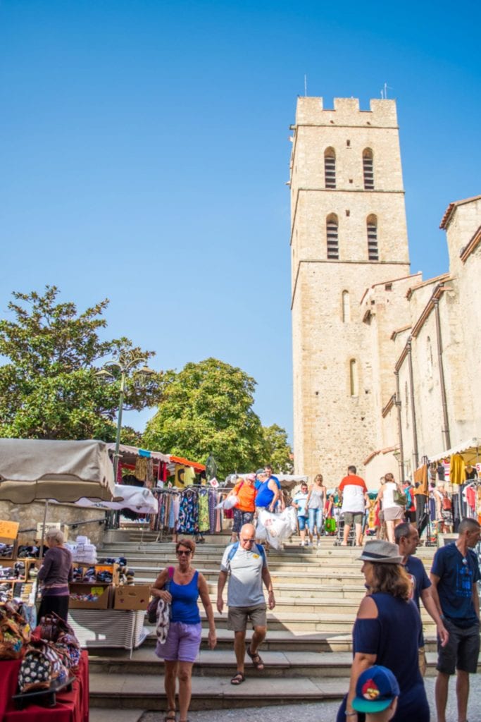 drukte tijdens de markt op een zomerse dag in het centrum van  Argelès-sur-Mer. Er lopen zomers geklede mensen op de trap naast een kerkgebouw.