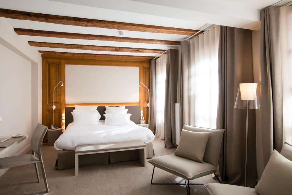 kamer met beige tinten, houten balken tegen het plafond, witte muren en wit beddengoed op een tweepersoonsbed in een kamer van het hotel dat is gevestigd in het Maison de Têtes