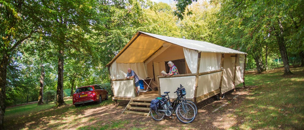 twee senioren op het overdekte terras van een glampingtent met beide een boekje voor hun hand. Voor de tent staan twee fietsen en er naast staat een rode auto met een Nederlands kenteken