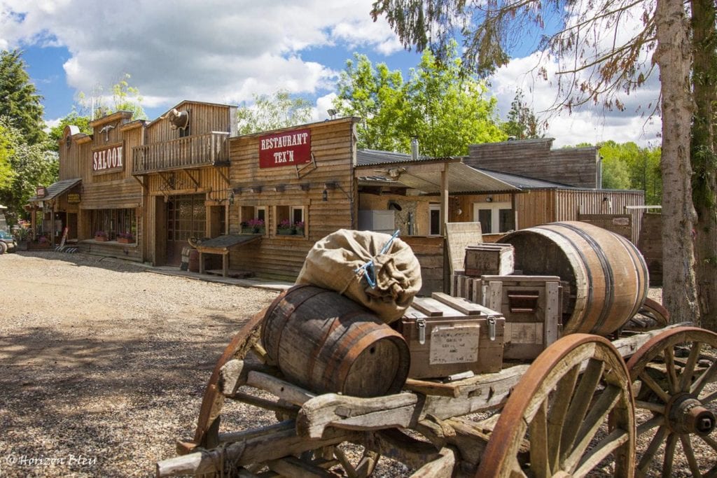 houten saloon met daarop de teksten 'saloon' en 'restaurant tex'n' in witte letters op een rode achtegrond, en een houten kar met tonnen, dozen en een zak op de voorgrond van de foto