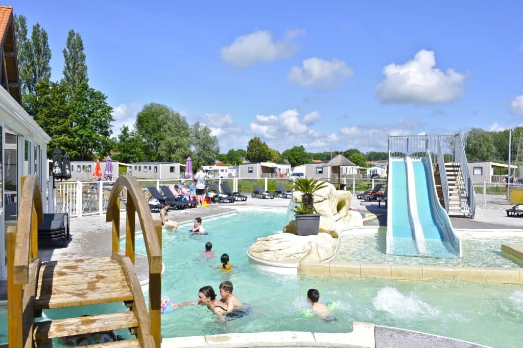 kinderen en twee vrouwen die zwemmen in het buitenzwembad van camping Domaine de la Roselière. Het zwembad heeft een bruggetej en twee glijbanen, en rondom staan grijze stacaravans.