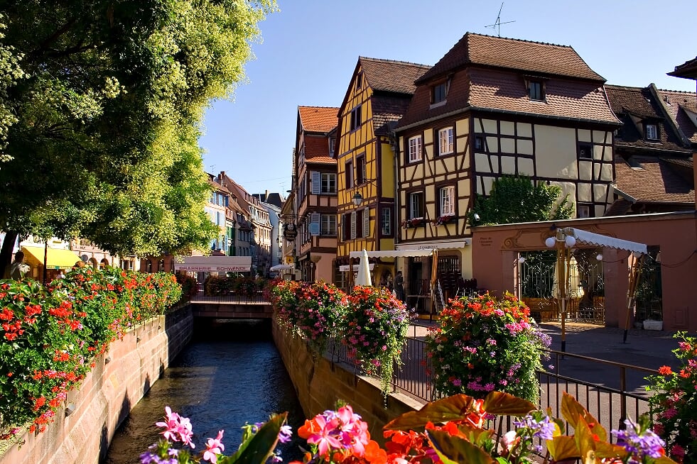 Straat op een zonnige dag in Colmar. Er loopt een kanaaltje door de straat met rondom bloembakken en aan de straat liggen gekleurde vakwerkhuizen