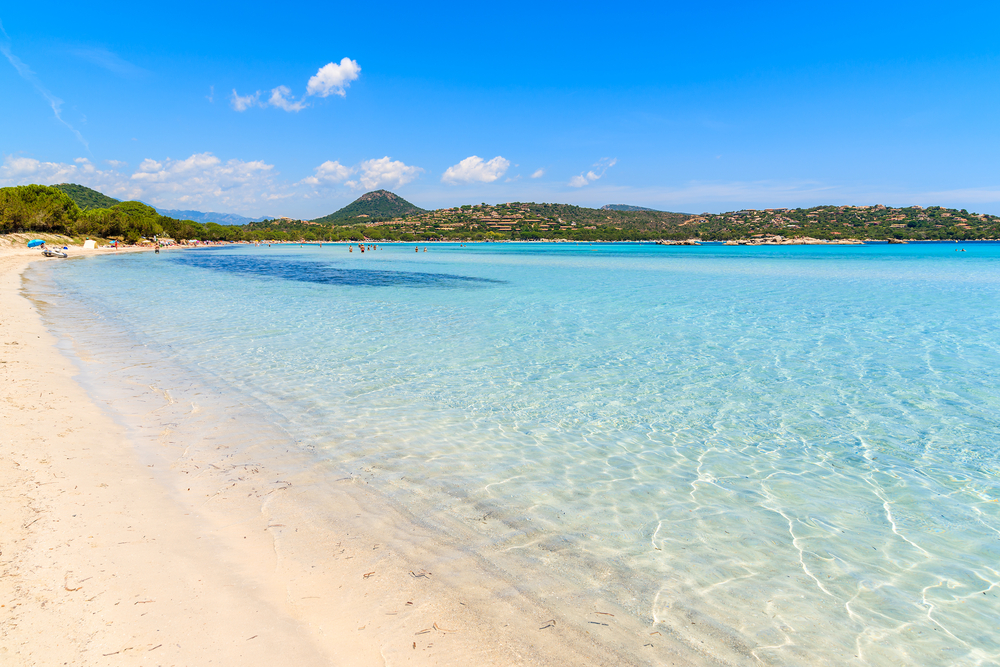 een heldere, lichtblauwe zee gelegen aan een lichtgekleurd zandstrand met in de verte mensen en groene heuvels op Corsica