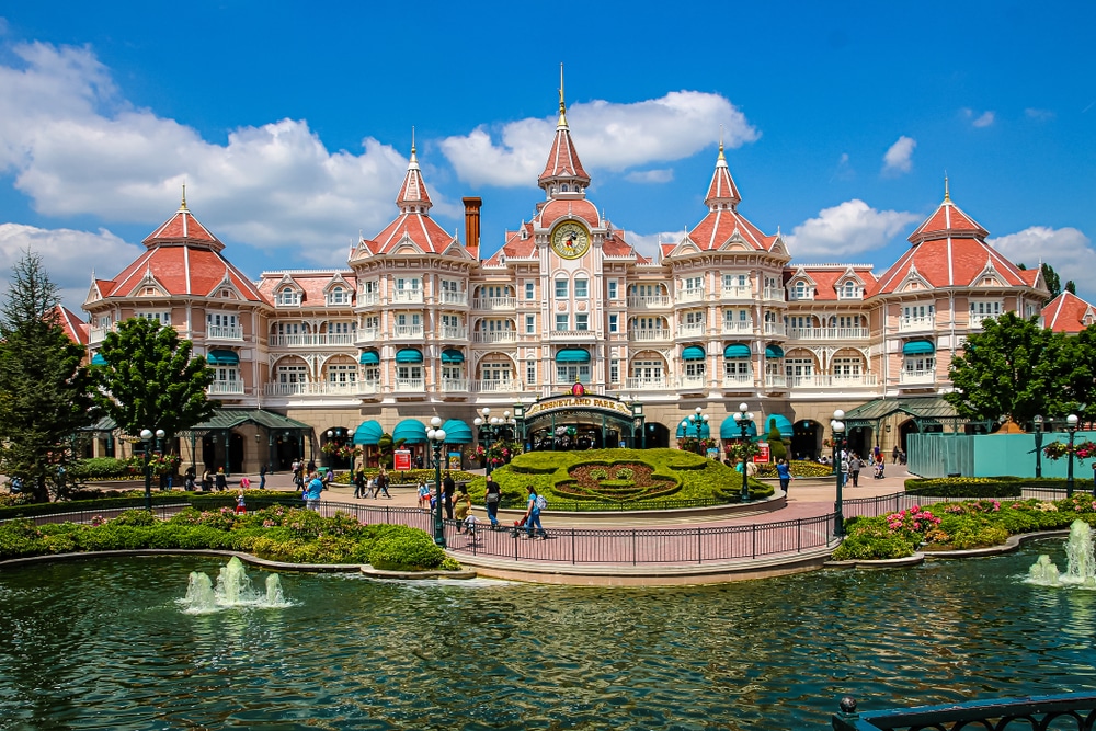 Het entreegebouw van Disneyland Parijs met roodgekleurde puntige daken gelegen aan water met fonteinen en een Mickey Mouse afbeelding gemaakt in gras.