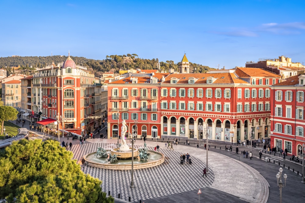 het plein Place Massena met zijn fontein en rondom hoge, oranje gebouwen. Er struinen een aantal mensen over het plein en door de straten