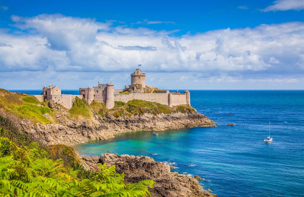Foto van een stenen fort gelegen aan de Bretonse kust omringd door begroeide rotsen en helderblauw water van de oceaan.