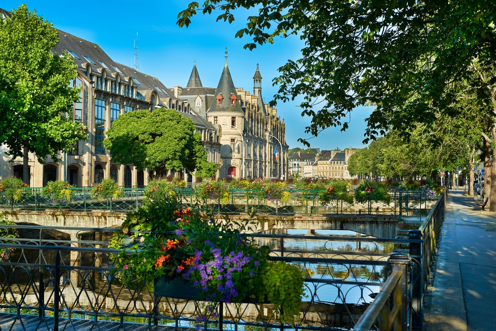 Foto van een kasteel omringd door wateren, andere gebouwen, mooie bruggen, kleurrijke bloemen en bomen in de stad Quimper.