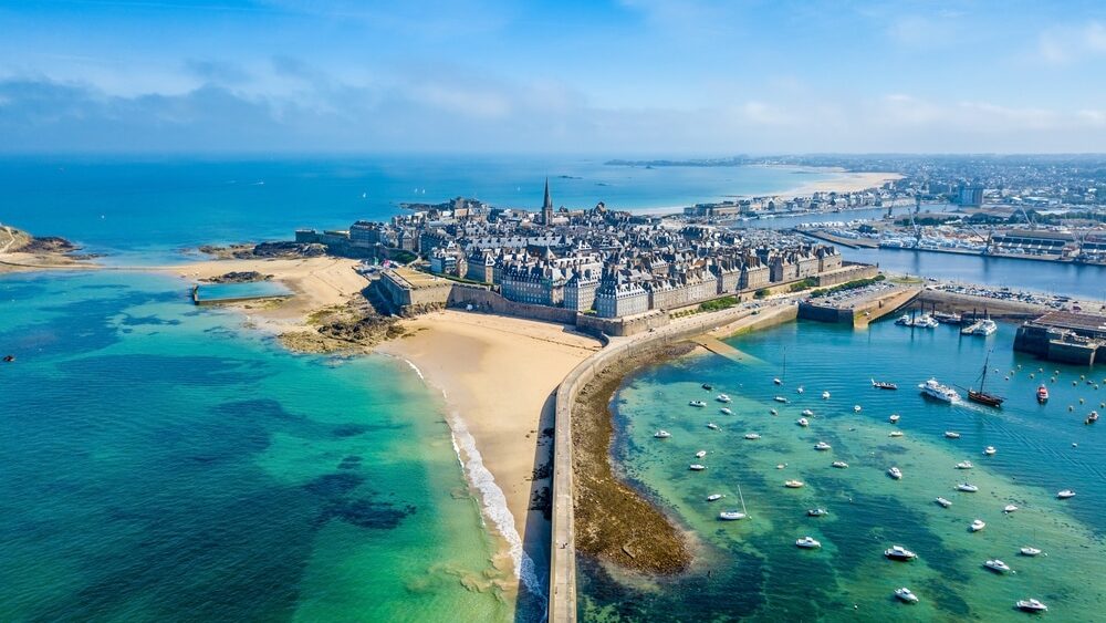 Luchtfoto van de kustplaats Saint-Malo. Een stad met veel gebouwen omringd door strand, helderblauwe wateren en witte bootjes.