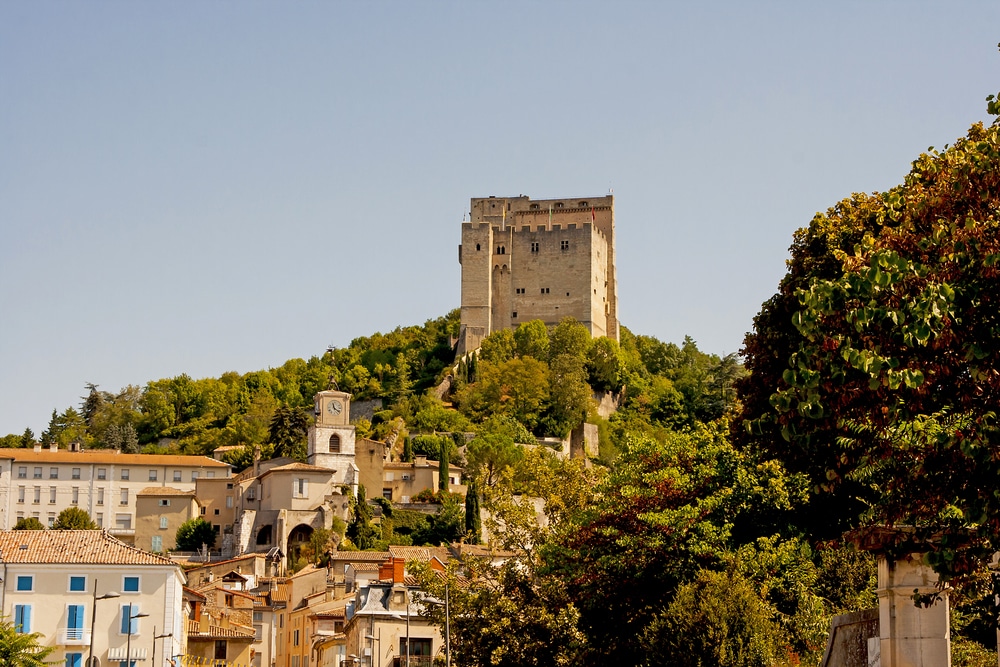 een toren op een met bomen begroeide berg die boven een stadje met huizen en kerktoren uitsteekt