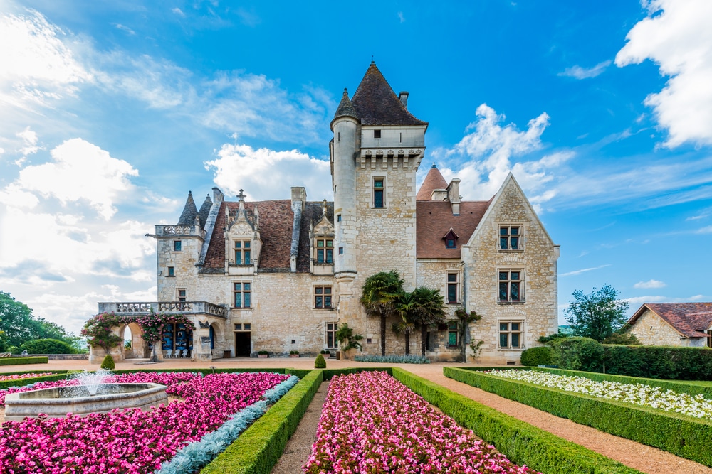 Schitterend kasteel met rode daken gelegen in een tuin met struiken, roze bloemen en een fontein.