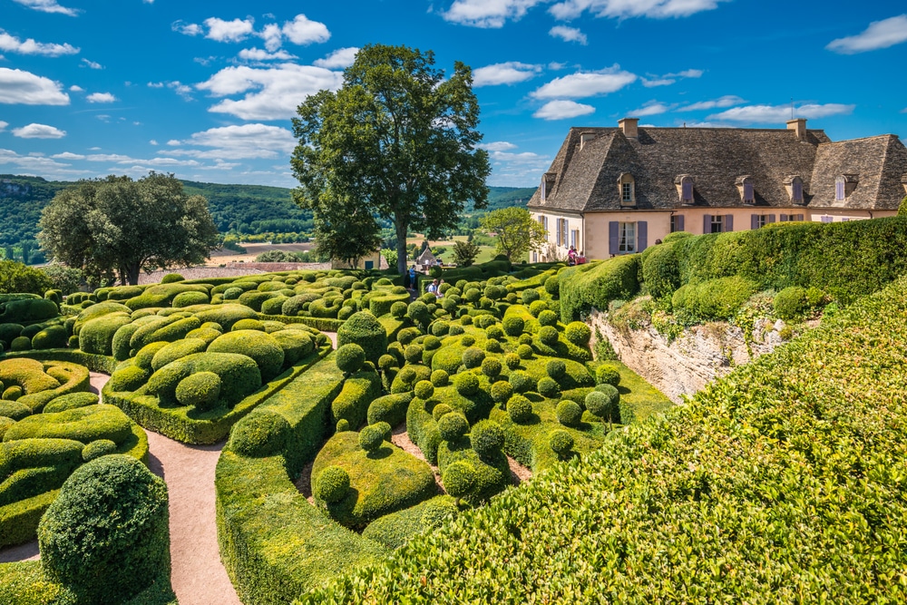 Een tuin met mooi gesnoeide struiken, vrijlopende paden, een boom en middeleeuws huisje.