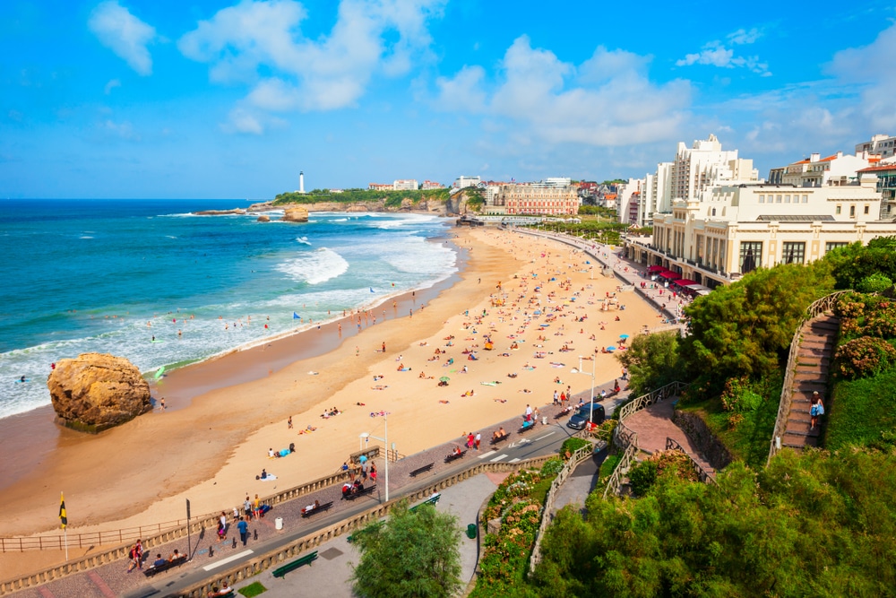 een druk en groot strand waarop strandlakens in alle kleuren liggen en daarachter een boulevard is met aan het einde een vuurtoren in Biarritz