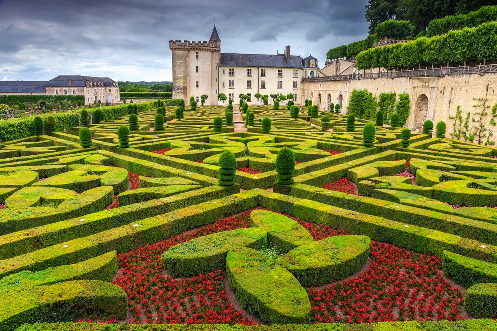 Mooi gevormde heggen en roodgekleurde bloemen grenzend aan een wit kasteel met zwarte daken.