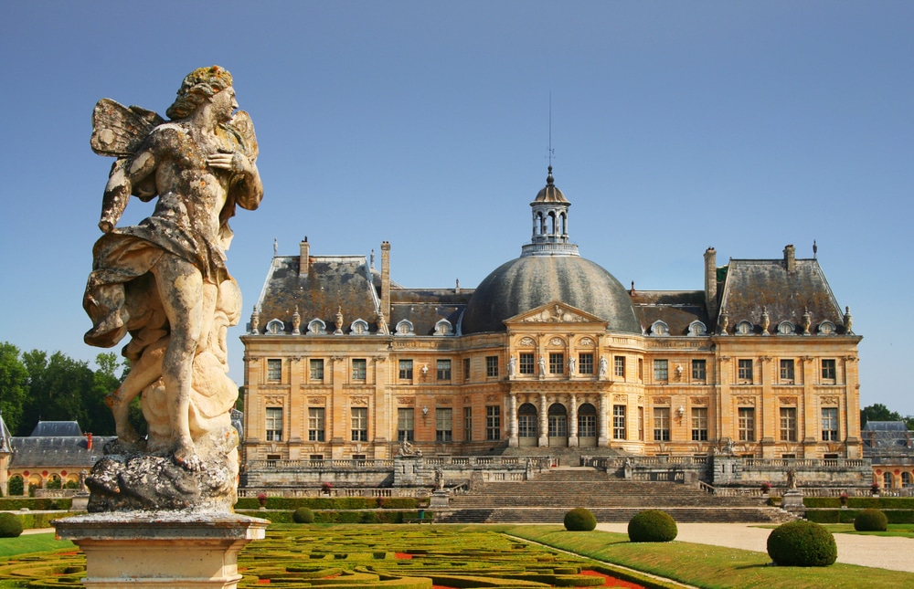 Het zandkleurige kasteel van Vaux-le-Vicomte met een zwart koepelvormig dak met daarop een toren. Gelegen in de kasteeltuin met mooi gevormde heggen en een stenen beeld van een engel.