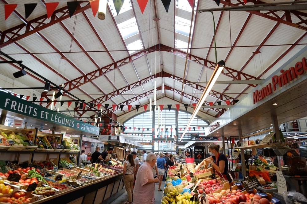 Binnenkant van een marktgebouw met daarin kraampjes met fruit, vlaggetjes en mensen die aan het rondkijken zijn.