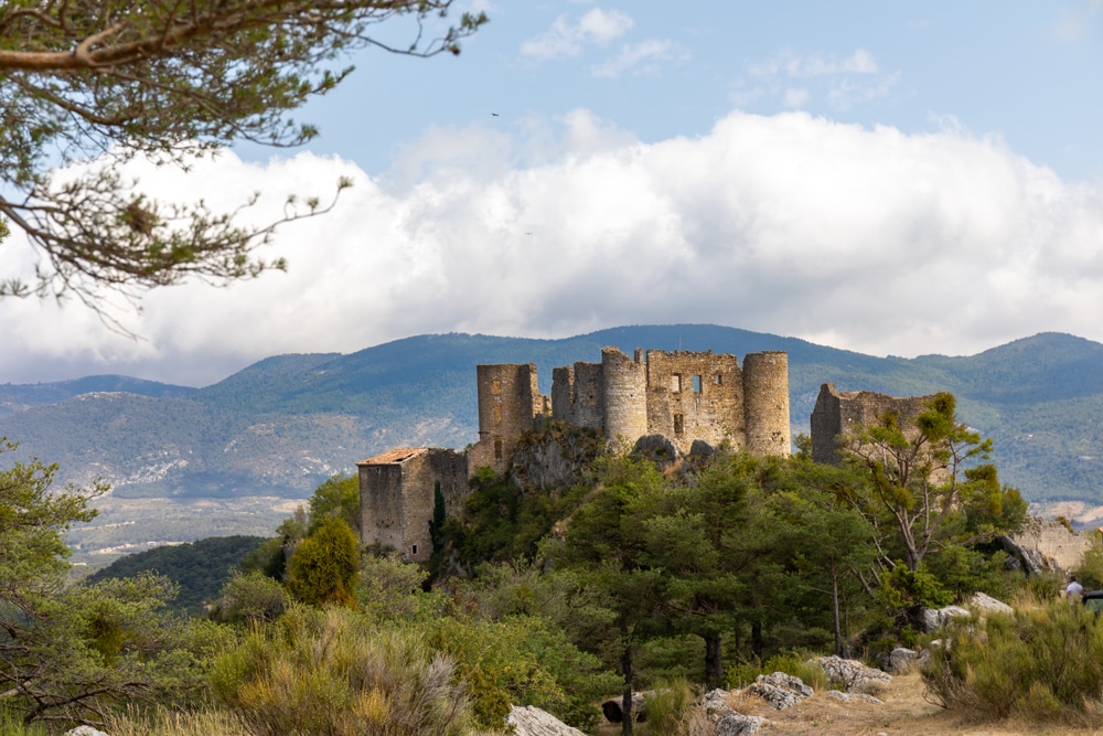 Stenen ruïnes van een oud kasteel gelegen hoog op een rots, omringd door groene bomen en met uitzicht op de omliggende bergen.