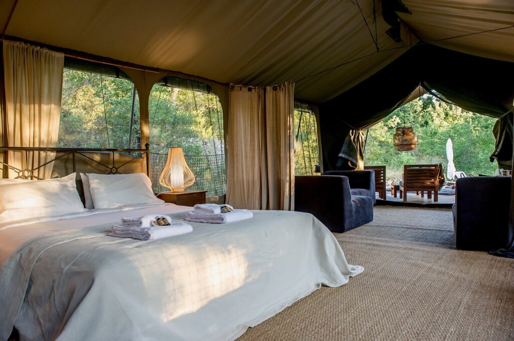 luxe safaritent met een strak opgemaakt tweepersoonsbed, loungstoelen en op de achtergrond het terras