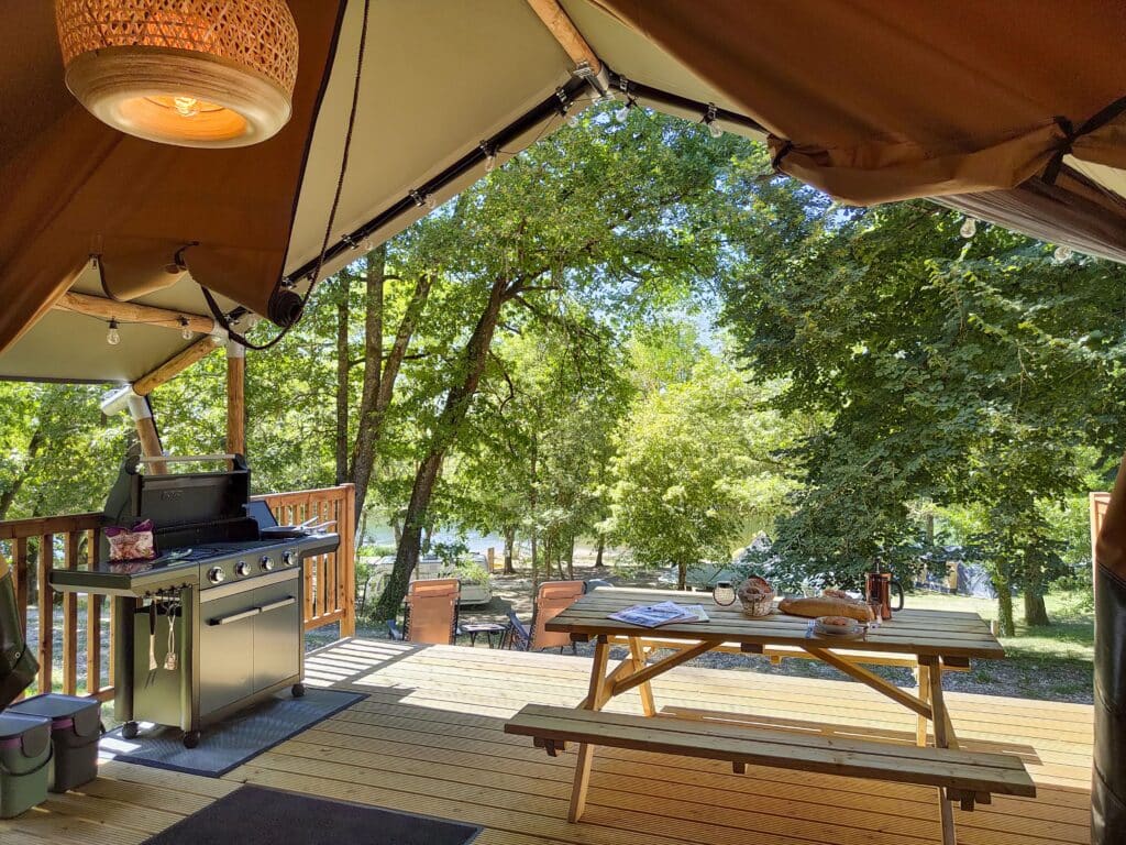 het terras van een safaritent op Camping Le Clos Bouyssac met een moderne barbecue en ene picknicktafel met brood en tijdschriften