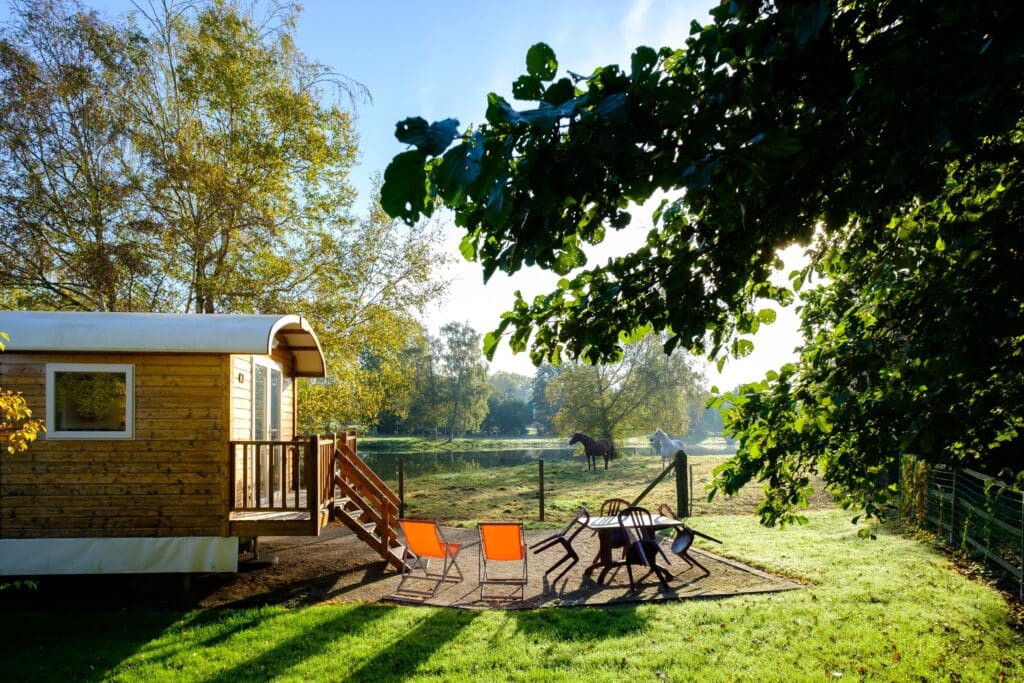 gesloten pipowagen met daarover een terras met twee oranje zonnestoeltjes en een tafel met vier stoelen op Domaine Du Lieu Dieu. Op de achtergond staan een wit en een zwart paard in een weiland