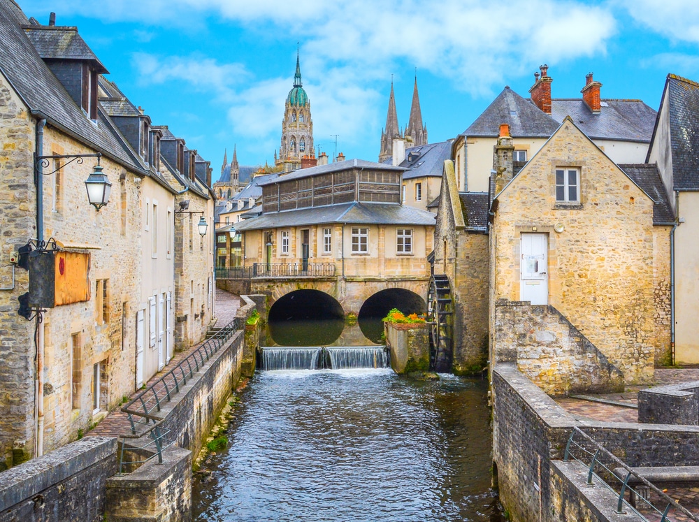 kanaaltje in het dorpje Bayeux met rondom huisjes en een bruggetje en op de achtergrond de torens van de kathedraal die boven de andere gebouwen uitsteken