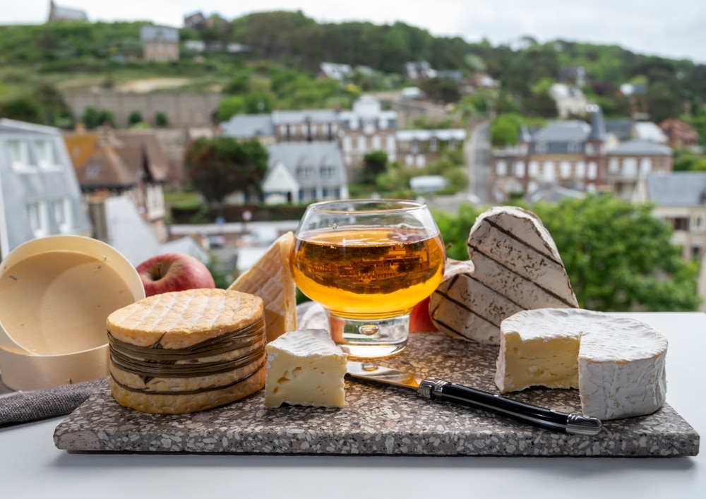 een glas met Calvados en rondom verschillende kazen op een plankje met op de achtergrond de het typische landschap en de huizen in de buurt van de Normandische kust