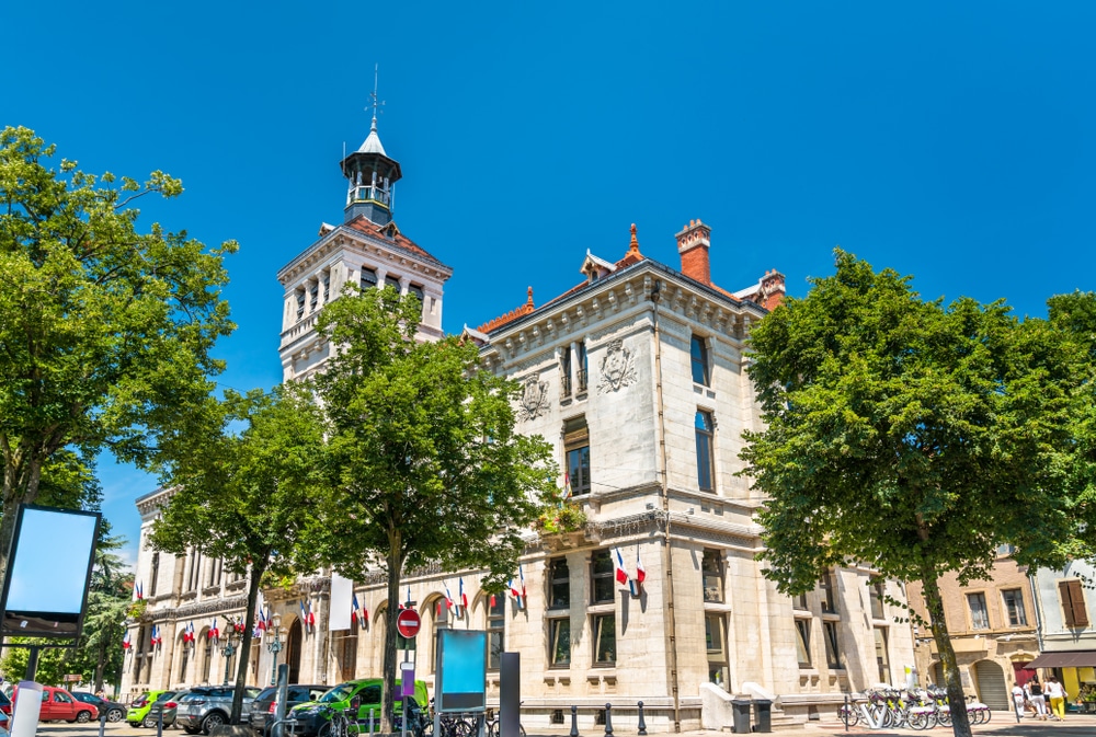 het stadhuis van Valence waar meer dan tien kleine Franse vlaggen aan de gevel hangen. Voor het stadhuis staan auto's en bomen en aan de zijkant deelfietsen.